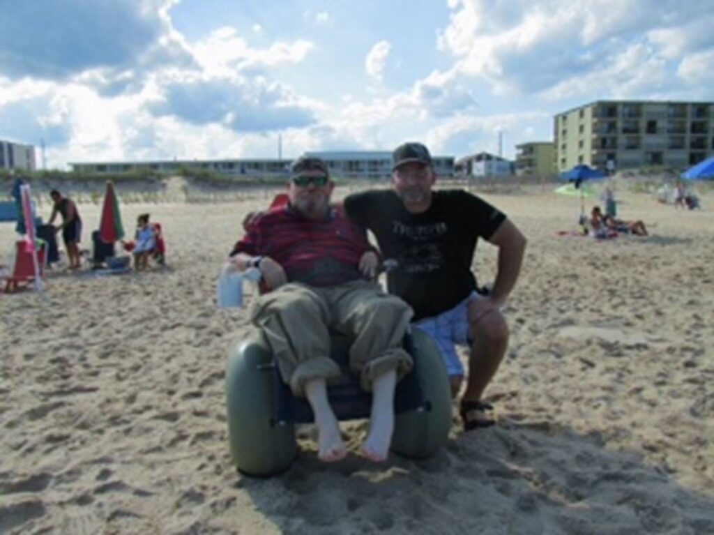 Jack Provencal and Rob Litchfield smiling on a sandy beach, one seated in a beach wheelchair.